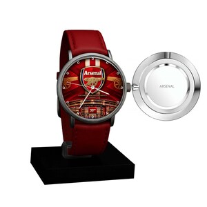 May include: A wristwatch with a red leather strap and a round silver case. The watch face features the Arsenal football club logo, stadium imagery, and the text Arsenal. A circular silver cover attached to the side displays the word Arsenal.