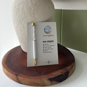 Op de afbeelding: Een witte balpen met een doorzichtige houder gevuld met kleine witte steentjes, bevestigd aan een rechthoekige kaart. De kaart bevat een illustratie van een maansteen, de tekst "moonstone", "new chapter" en een lijst met eigenschappen.
