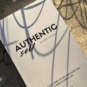 May include: A light grey guided journal with black text on the cover that reads AUTHENTIC self GUIDED JOURNAL and YOUR JOURNEY OF SELF-DISCOVERY AND A MORE AUTHENTIC LIFE. The cover features a matte finish with shadows cast across the surface.