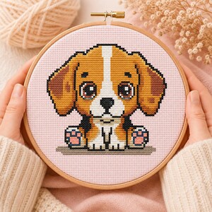 Pode incluir: Um projeto de bordado em ponto cruz num bastidor de madeira redondo com um cachorrinho de desenho animado com olhos castanhos grandes, uma risca branca no rosto e pelo bege e branco num fundo de tecido rosa claro com padrão de grelha.