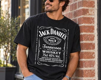 May include: A black short-sleeve t-shirt featuring a white graphic print of the Jack Daniel's Old No. 7 Tennessee Sour Mash Whiskey label, including the text 75cl 40% Vol. and the Jack Daniel Distillery Lynchburg, Tennessee 37352 U.S.A. address.