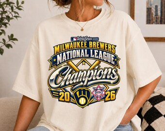May include: An off-white short-sleeve t-shirt featuring a graphic for the Milwaukee Brewers 2026 National League Champions. The design includes the team name, baseball bats, a baseball, the year 2026, and the MLB World Series logo.