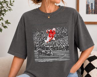 May include: A charcoal grey short-sleeve t-shirt featuring a graphic print of a football player in a red jersey leaping over another player on a field. The design includes a stadium crowd background and the handwritten-style text DM in white.
