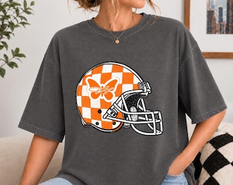 May include: A charcoal grey short-sleeve t-shirt featuring a graphic of a football helmet with an orange and white chequered pattern. The helmet includes a white butterfly design on the side and a white face mask. The shirt has a crew neckline.