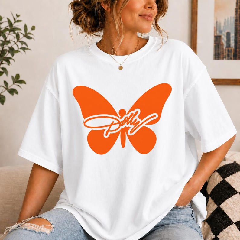 May include: A white short-sleeve t-shirt featuring a large, solid orange butterfly graphic centered on the chest. The word Dolly is written in a white cursive script font across the middle of the butterfly wings. The shirt has a crew neckline.