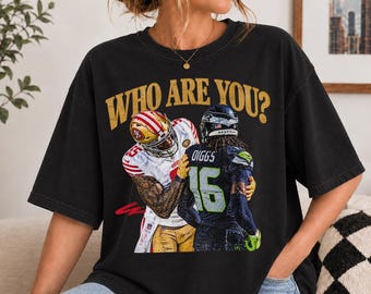 May include: A black short-sleeve t-shirt featuring a graphic print of two football players in a confrontation. The text WHO ARE YOU? is printed in gold above the players. One player wears a white jersey and the other wears a dark jersey with DIGGS 16.