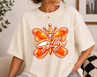 May include: An off-white short-sleeve t-shirt featuring a large, glossy orange butterfly graphic centred on the chest. The butterfly is topped with a small orange crown and displays the word Polly in a white cursive script across its wings.