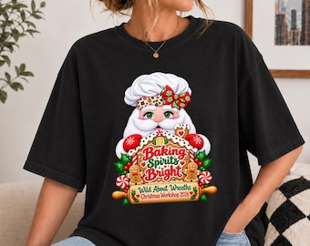 May include: A black t-shirt featuring a graphic of a chef Santa Claus wearing a white hat with a red and green bow. The design includes a gingerbread house, candy canes, holly, and the text Baking Spirits Bright Wild About Wreaths Christmas Workshop 2026.