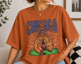 May include: A rust-coloured short-sleeve t-shirt featuring a graphic print with the word CHICAGO in blue letters above a football player riding a bear, surrounded by orange flames and a green patch of grass at the bottom of the design.