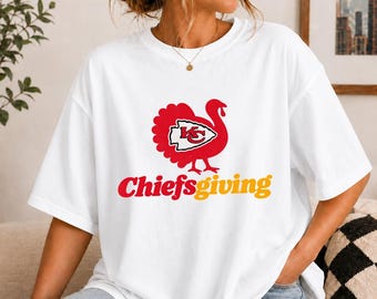 May include: A white short-sleeved t-shirt featuring a red graphic of a turkey with a Kansas City Chiefs arrowhead logo on its body. Below the turkey, the text Chiefs-giving is printed in red and yellow lettering on the front.