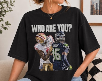 May include: A black t-shirt featuring the text WHO ARE YOU? above a graphic of two football players. One player wears a white and gold 49ers uniform, and the other wears a navy blue and neon green Seahawks uniform with the name DIGGS on the back.