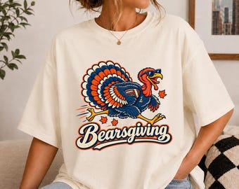 May include: An off-white short-sleeve t-shirt featuring a graphic of a turkey wearing sunglasses and holding a football. The turkey has blue and orange feathers. Below the graphic, the text Bearsgiving is printed in a bold, dark blue font.