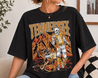 May include: A black short-sleeve t-shirt featuring a graphic print with the word TENNESSEE in large tan letters. The design includes a skeleton football player in a yellow jersey, a dog, mountains, and flames in shades of orange and yellow.