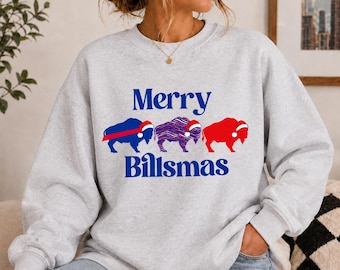 May include: A light grey crewneck sweatshirt featuring the blue text Merry Billsmas. Between the words are three buffalo silhouettes, each wearing a red and white Santa hat. The first buffalo is solid blue, the second is blue and red striped, and the third is solid red.