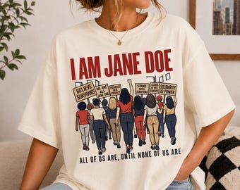 May include: An off-white t-shirt featuring a graphic of a group of people holding protest signs. The text reads I AM JANE DOE at the top and ALL OF US ARE, UNTIL NONE OF US ARE at the bottom. The signs include phrases like BELIEVE SURVIVORS.
