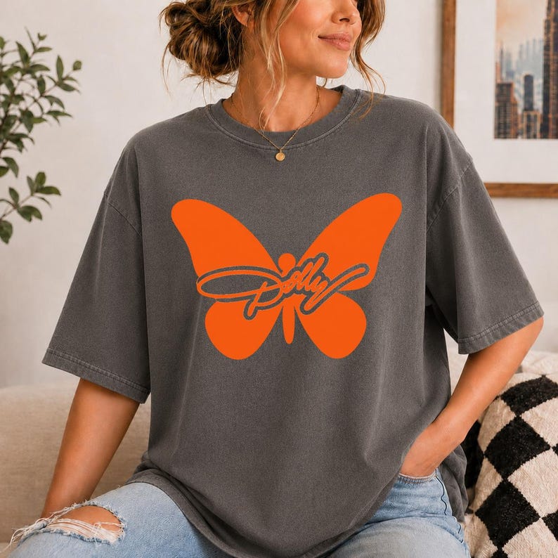 May include: A charcoal gray short-sleeve t-shirt featuring a large, solid orange butterfly graphic centered on the chest. Overlaid on the butterfly is the word Dolly written in a dark gray cursive script font with a thin orange outline.