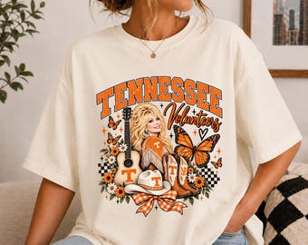 May include: An off-white short-sleeve t-shirt featuring a graphic print with the text TENNESSEE Volunteers. The design includes a blonde woman, an acoustic guitar, a cowboy hat, cowboy boots, monarch butterflies, and orange flowers.