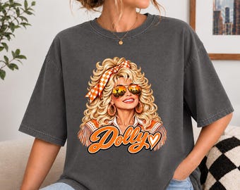 May include: A charcoal grey short-sleeve t-shirt featuring a graphic print of a blonde person with voluminous hair, wearing orange-tinted sunglasses and an orange chequered headband. The word Dolly is printed in orange script below the graphic.