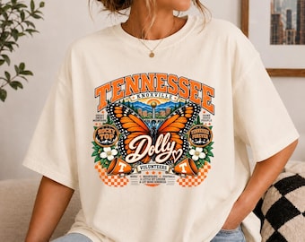 May include: A cream-coloured t-shirt featuring a large graphic print with the text TENNESSEE, KNOXVILLE, and Dolly. The design includes a monarch butterfly, orange megaphones, white flowers, and a checkered pattern in orange and white colours.