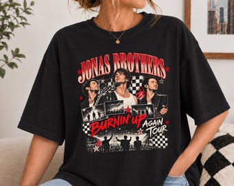 May include: A black short-sleeve t-shirt featuring a graphic print of the Jonas Brothers. The design includes the text JONAS BROTHERS, BURNIN' UP, and AGAIN TOUR, along with concert imagery, red stars, and black and white checkered patterns.