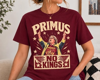 May include: A maroon short-sleeve t-shirt featuring a graphic print of a person with glasses and a yellow jacket. The text reads PRIMUS at the top and NO KINGS at the bottom, surrounded by star accents and radiating lines in a vintage style.