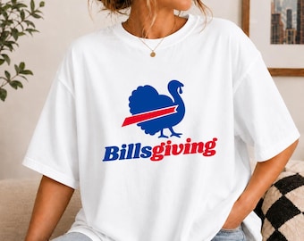 May include: A white short-sleeve t-shirt featuring a blue graphic of a turkey silhouette with a red horizontal stripe across its center. Below the graphic, the text Billsgiving is printed in blue and red lettering on the front of the shirt.