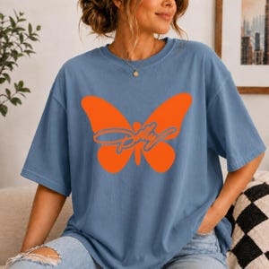 May include: A dusty blue short-sleeve t-shirt featuring a large, solid orange butterfly graphic centered on the chest. Overlaid on the butterfly is the word Dolly written in a cursive, blue-outlined font. The shirt has a crew neckline.