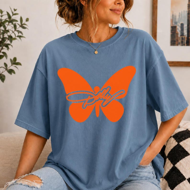 May include: A dusty blue short-sleeve t-shirt featuring a large, solid orange butterfly graphic centered on the chest. Overlaid on the butterfly is the word Dolly written in a cursive, blue-outlined font. The shirt has a crew neckline.