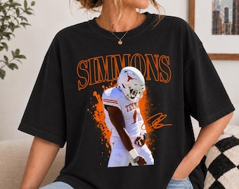 May include: A black short-sleeve t-shirt featuring a graphic of a football player in a white uniform with the name SIMMONS in large orange letters above. The design includes orange paint splatter effects and a signature in orange ink.