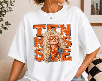 May include: A white short-sleeved t-shirt featuring a graphic print of a blonde woman with voluminous hair wearing orange star-patterned sunglasses and a matching orange bandana. The text TENNESSEE is printed in large, distressed orange block letters.