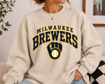 May include: A cream-colored crewneck sweatshirt featuring the text MILWAUKEE BREWERS in dark blue arched lettering above a dark blue and yellow baseball glove icon containing a white baseball graphic on the chest.