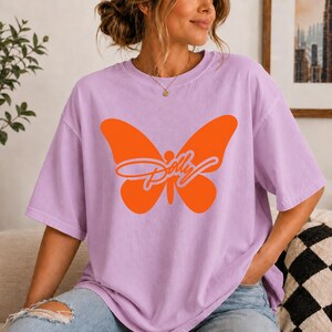 May include: A light purple short-sleeve t-shirt featuring a large, solid orange butterfly graphic centered on the chest. Overlaid on the butterfly is the word Dolly written in a cursive, light purple script font with a thin white outline.