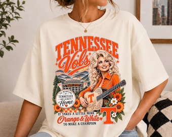 May include: An off-white t-shirt featuring a graphic print of a blonde woman playing an acoustic guitar with a large orange T logo. The design includes text reading Tennessee Vols, The Mountains of Tennessee Are Always Home, and Orange & White.