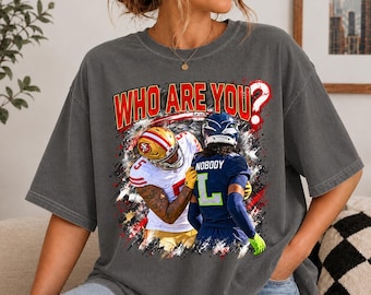 May include: A charcoal grey short-sleeve t-shirt featuring a graphic print of two football players in opposing uniforms. The text WHO ARE YOU? appears in red above the players, and the word NOBODY is printed on the back of the dark blue jersey.