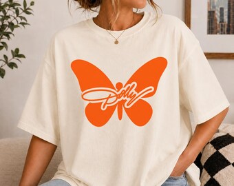 May include: An off-white short-sleeve t-shirt featuring a large, solid orange butterfly graphic centred on the chest. The word Dolly is written in a white cursive script font across the middle of the butterfly wings. The shirt has a crew neckline.