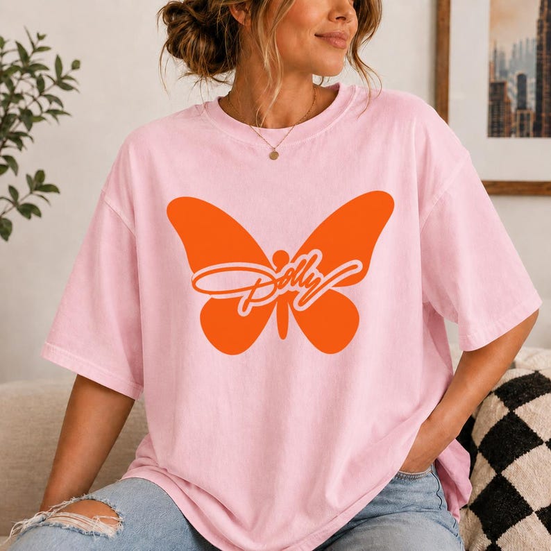 May include: A light pink short-sleeve t-shirt featuring a large, solid orange butterfly graphic centered on the chest. Overlaid on the butterfly is the word Dolly written in a white cursive script font. The shirt has a crew neckline.