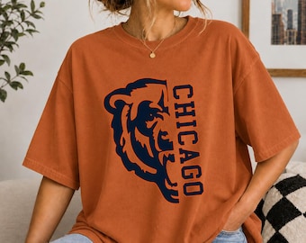 May include: A burnt orange short-sleeve t-shirt featuring a dark navy blue graphic on the front. The graphic displays the left half of a bear's face alongside the vertical text CHICAGO in a bold, blocky, dark navy blue font.