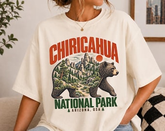 May include: An off-white t-shirt featuring a graphic of a bear with a forest and mountain landscape inside its silhouette. The text reads CHIRICAHUA NATIONAL PARK ARIZONA, USA, with orange and green lettering and a small sun icon above the bear.