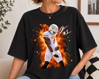 May include: A black t-shirt featuring a graphic of a football player in a white uniform with the word TEXAS and the number 1, surrounded by orange flames and splatter effects. A white signature and the text #1 are printed above the player.
