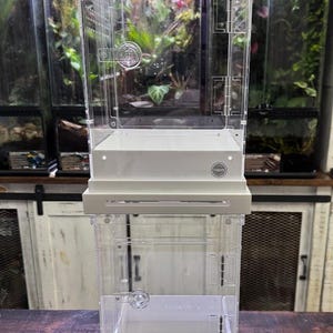 May include: A vertical, clear acrylic terrarium enclosure featuring a white base and a hinged front door with a circular latch. The structure includes a Thrive brand logo on the front panel and a ventilated top section for airflow and habitat maintenance.