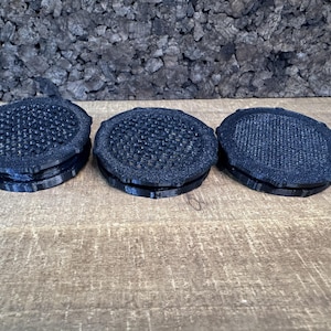 May include: Three circular black coasters with a textured, grid-like pattern on the top surface. Each coaster features a slightly raised, scalloped edge design and a solid black base, presenting a uniform appearance across all three individual items.