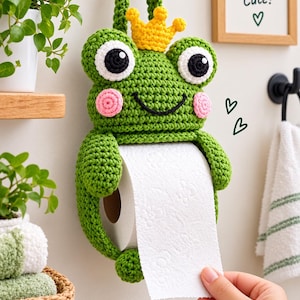Puede incluir: Un soporte para papel higiénico de ganchillo verde con forma de rana, que incluye una corona amarilla, ojos grandes blancos y negros y mejillas rosas. La rana tiene un lazo para colgar y sostiene un rollo de papel higiénico blanco.