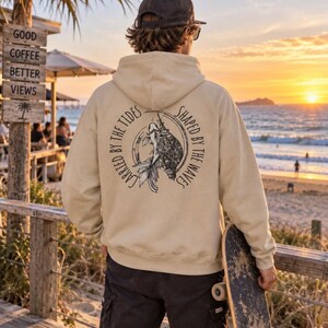 May include: A beige hooded sweatshirt featuring a black circular graphic on the back. The design depicts a fish and includes the text "CARRIED BY THE TIDES SHAPED BY THE WAVES" written in a circular pattern around the central illustration.