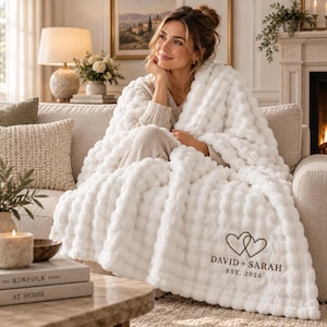 May include: A white plush textured throw blanket featuring a repeating pom-pom pattern. The blanket is embroidered with two interlocking heart outlines above the text "DAVID + SARAH" and "EST. 2026" in a dark brown, clean sans-serif font.