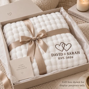 Peut inclure: Une couverture blanche texturée à motif de bulles, enveloppée d'un ruban de satin couleur champagne noué en nœud. La couverture présente un motif brodé marron foncé de deux cœurs entrelacés avec le texte DAVID + SARAH EST. 2026.
