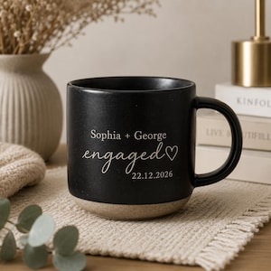May include: A black ceramic mug with a light-coloured base featuring the white engraved text Sophia + George, the word engaged in a script font, a heart symbol, and the date 22.12.2026 centred on the front of the mug with a side handle.