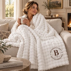 May include: A white plush textured throw blanket featuring a circular brown embroidered design with the text THE BROWN FAMILY ESTABLISHED 2026 and a central letter B with a small heart symbol, draped over a sofa while holding a ceramic mug.