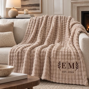 May include: A beige, textured, waffle-knit throw blanket draped over a sofa. The blanket features a dark brown embroidered monogram with the letters E and M separated by a vertical line, flanked by leaf sprigs, and the text EST. 2026 below.
