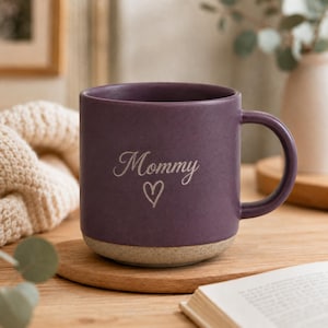 May include: A matte purple ceramic mug with a rounded handle and a textured beige base. The front of the mug features the word Mommy in a cursive script font, positioned directly above a simple, hand-drawn outline of a heart shape.