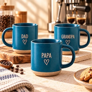 May include: Three teal ceramic mugs with a speckled beige base. Each mug features a white heart outline and text: one reads DAD, one reads GRANDPA, and one reads PAPA. The mugs have a smooth finish and a sturdy handle on the right side.